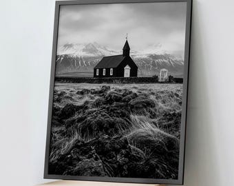 May include: A black and white framed wall art print featuring a dark church with a steeple, situated in a field of rocky terrain and tall grass, with snow-capped mountains under a cloudy sky in the background, presented in a thin black frame.