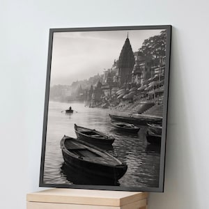 May include: A black and white framed wall art print depicting a river scene with several wooden rowboats floating on the water. In the background, stone steps lead up to a series of buildings and a tall temple spire under a hazy, overcast sky.
