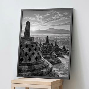 Può includere: Una stampa artistica da parete incorniciata in bianco e nero che mostra una vista paesaggistica degli stupa del tempio di Borobudur con ritagli a forma di diamante. La scena cattura un'alba su montagne lontane e una valle nebbiosa.