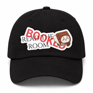 Könnte beinhalten: Eine schwarze Baseballkappe mit einem grafischen Design, das den roten Schriftzug BOOKED und den schwarzen Schriftzug READ THE ROOM zeigt. Rechts neben dem Text befindet sich eine Cartoon-Illustration eines braunen Buches mit Gesicht, weißen Flügeln und dem Titel The Room auf dem Einband.
