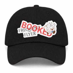 Könnte beinhalten: Eine schwarze Baseballkappe mit einer Grafik, die den roten Schriftzug BOOKED und den schwarzen Schriftzug FREQUENT FLYER zeigt. Neben dem Text befindet sich eine Comicfigur, die einem weißen Reiseadapter mit Gesicht, blauer Fliege und braunen Füßen ähnelt.
