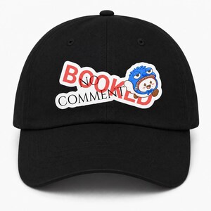 Könnte beinhalten: Eine schwarze Baseballkappe mit einem grafischen Design, das den roten Schriftzug BOOKED und den schwarzen Schriftzug NO COMMENT zeigt. Neben dem Text befindet sich eine Zeichentrickfigur mit blauem Fell und weißem Gesicht sowie blauer Fliege.