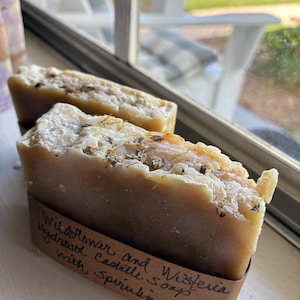 Può includere: Due barre rettangolari di sapone beige con cime strutturate incastonate con pezzi botanici essiccati. Una fascia di carta marrone avvolge la parte anteriore del sapone, con il testo scritto a mano Wildflower and Wisteria Hydrosol Castile Soap with Spirulina.