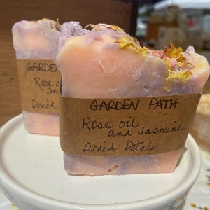 Può includere: Due saponette rettangolari fatte a mano con una tavolozza di colori marmorizzati pesca e lavanda. Ogni saponetta è avvolta in un'etichetta di carta marrone con il testo scritto a mano GARDEN PATH, Rose oil and Jasmine, Dried Petals.