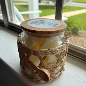 May include: A clear glass jar filled with white wax melts, encased in a woven light brown wicker sleeve. The jar features a wooden lid with a circular sticker displaying the text Yellow Flower Farm and a small wooden spoon attached to the side.