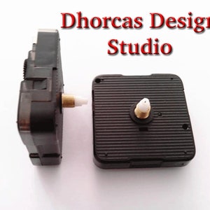 May include: Two black clock movements with gold-colored threaded shafts and white plastic inserts. The clock movements are next to each other on a white surface. The text "Dhorcas Design Studio" is in red at the top of the image.
