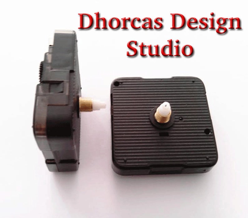 May include: Two black clock movements with gold-colored threaded shafts and white plastic inserts. The clock movements are next to each other on a white surface. The text "Dhorcas Design Studio" is in red at the top of the image.