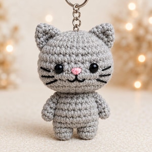 May include: A gray crocheted cat keychain featuring a round head with pointed ears, black plastic bead eyes, a small pink stitched nose, and black embroidered whiskers. The body is compact with short arms and legs, attached to a silver metal ring.