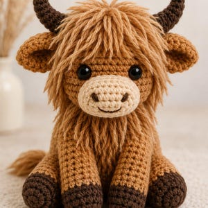 May include: A crocheted Highland cow stuffed animal with shaggy light brown yarn fur, a cream-colored muzzle, dark brown horns, and dark brown hooves. The toy features large black safety eyes and a stitched mouth, sitting in an upright position.