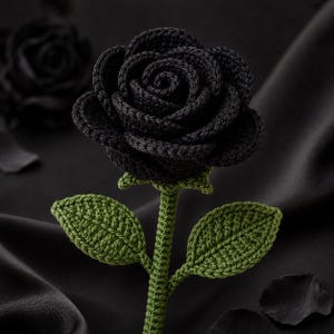 May include: A handcrafted crochet rose featuring a deep black bloom with tightly layered petals. The stem and two pointed leaves are worked in a textured olive green yarn, showcasing intricate stitch detail throughout the entire floral structure.
