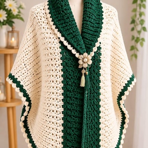 May include: A crochet poncho featuring alternating vertical panels of cream and dark green yarn. The garment has a wide dark green ribbed collar, a scalloped cream border, and a decorative metallic flower brooch with a pearl and green tassel.