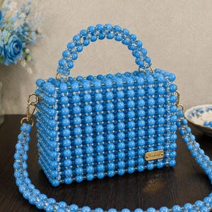 Könnte beinhalten: Eine rechteckige Handtasche aus leuchtend blauen, kugelförmigen Perlen in einem Gittermuster. Die Tasche verfügt über einen passenden Perlen-Tragegriff und einen abnehmbaren Perlen-Schulterriemen mit goldfarbenen Metallbeschlägen und einem kleinen rechteckigen Schild mit der Aufschrift Handmade.