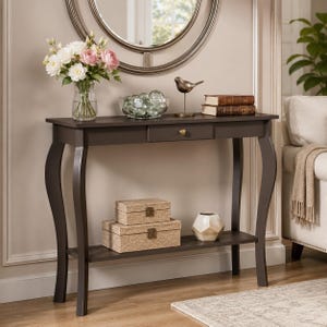 May include: A dark brown wooden console table with curved legs, a single drawer with a small round knob, and a lower shelf. The table holds a glass vase with white and pink flowers, a glass bowl with decorative spheres, a bird figurine, and books.