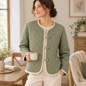 Peut inclure: Une veste matelassée vert sauge avec un motif floral répétitif blanc et foncé, une encolure ronde et des manches longues. La veste présente un liseré de couleur crème sur les bords et se ferme à l'avant avec trois boutons vert sauge assortis.