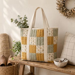 Op de afbeelding: Een gewatteerde tote bag met een patchworkontwerp van mosterdgele vierkanten, saliegroene ruitjes en crèmekleurige vierkanten met een klein bloemenpatroon. De tas heeft twee lange, crèmekleurige stoffen hengsels aan de voorzijde.