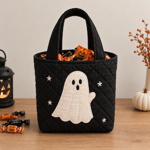 Könnte beinhalten: Eine schwarze, gesteppte Stofftasche mit einer weißen Geister-Applikation auf der Vorderseite, umgeben von kleinen gestickten weißen Sternen. Die Tasche hat zwei Tragegriffe und ist mit orangefarbenen und schwarzen Bonbons gefüllt.