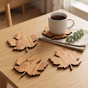 Könnte beinhalten: Drei hölzerne Untersetzer in Form von Ahornblättern mit sichtbarer Holzmaserung. Ein Untersetzer trägt eine weiße Keramiktasse mit einem dunklen, dampfenden Getränk, die auf einer gefalteten hellen Stoffserviette neben einem kleinen grünen Zweig steht.