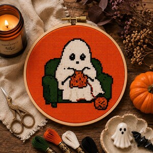 May include: A circular wooden embroidery hoop containing a cross-stitch pattern of a white ghost sitting in a dark green armchair, knitting with orange yarn against a bright orange background. The ghost has black eyes and a small black mouth.