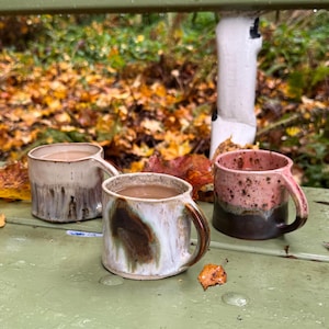 May include: Three ceramic mugs with handles, each featuring a unique glaze finish. One mug is cream with brown drips, another is white with a large brown and green splash, and the third is pink with dark brown speckles and a dark brown base.