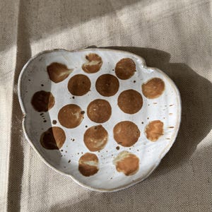 Könnte beinhalten: Ein handgeformter Keramikteller mit unregelmäßiger, organischer Form. Die Oberfläche ist cremeweiß mit glänzendem Finish und zeigt ein Muster aus verstreuten, soliden braunen kreisförmigen Punkten unterschiedlicher Größe sowie kleinen Sprenkeln.