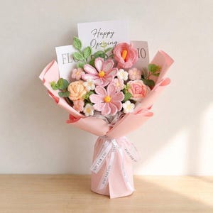 Può includere: Un bouquet di fiori all'uncinetto nei toni del rosa, pesca e bianco, con foglie verdi e un anthurium rosa centrale. La composizione è avvolta in carta rosa chiaro, legata con un nastro bianco con la scritta "JUST FOR YOU" e include un biglietto con "Happy Opening".