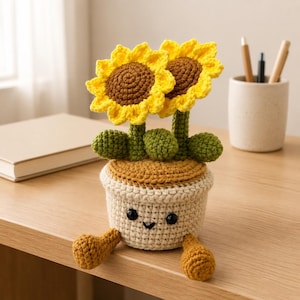 Op de afbeelding: Een gehaakte zonnebloemplant in een beige pot met een lachend gezicht, bestaande uit twee gele bloemen met bruine harten, groene stengels en bladeren. De pot heeft twee kleine bungelende benen, zwarte plastic ogen en een geborduurde mond.