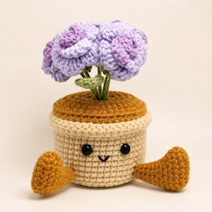 May include: A crocheted potted plant featuring a light beige cylindrical pot with a dark brown rim and two outstretched brown feet. The pot has two black bead eyes and a small stitched smile. A cluster of light purple crocheted flowers sits on top.