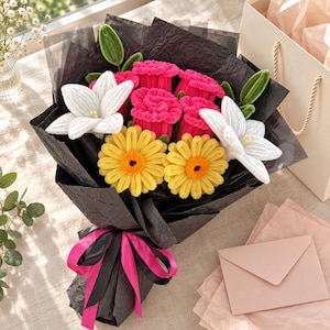 May include: A bouquet of crocheted flowers featuring bright pink roses, yellow daisies with orange centres, and white lilies with green buds. The arrangement is wrapped in black crinkled paper and tied with a vibrant pink and black ribbon.