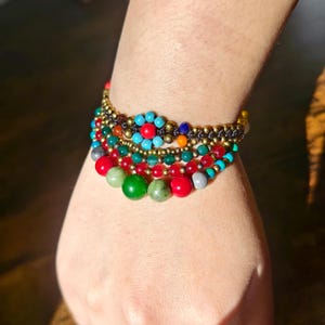 May include: A multi-layered bracelet featuring a variety of round beads in shades of red, green, turquoise, and metallic gold. The design includes a central flower-shaped motif made of turquoise and red beads, strung on a dark cord base.