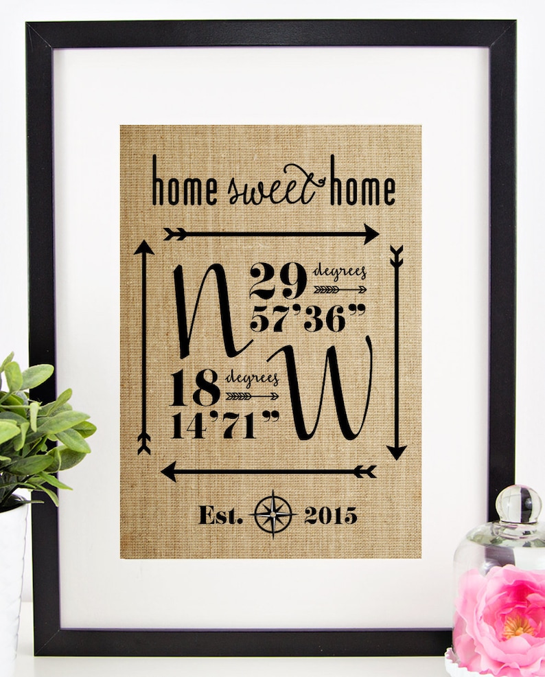 May include: A framed print on burlap fabric with the text "home sweet home" and coordinates: N 29 degrees 57'36" and W 18 degrees 14'71". The print also includes a compass and the text "Est. 2015".