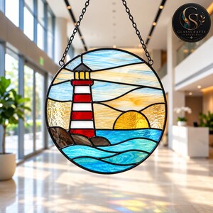Op de afbeelding: Een ronde glas-in-lood zonnevanger met een vuurtoren met rode en witte horizontale strepen, een gele zon en blauwe oceaangolven. Het stuk is omlijst in zwart metaal en hangt aan een zwarte ketting, met een afbeelding van een kustlandschap.