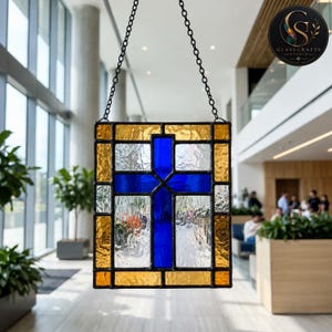 May include: A rectangular stained glass panel featuring a central blue cross set against a textured clear glass background, framed by amber-colored glass borders. The piece is suspended by a black metal chain and features a dark metal solder frame.