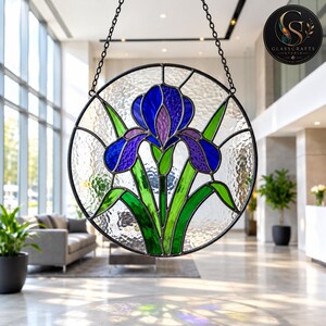 May include: A circular stained glass suncatcher featuring a central iris flower with deep blue and purple petals, surrounded by vibrant green leaves. The background consists of clear, textured glass panels held together by dark metal lead lines.