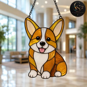 May include: A stained glass suncatcher shaped like a Corgi dog with amber, white, and black glass pieces. The dog has large pointed ears, a white chest, and a pink tongue. It is suspended by a black metal chain against a blurred interior background.