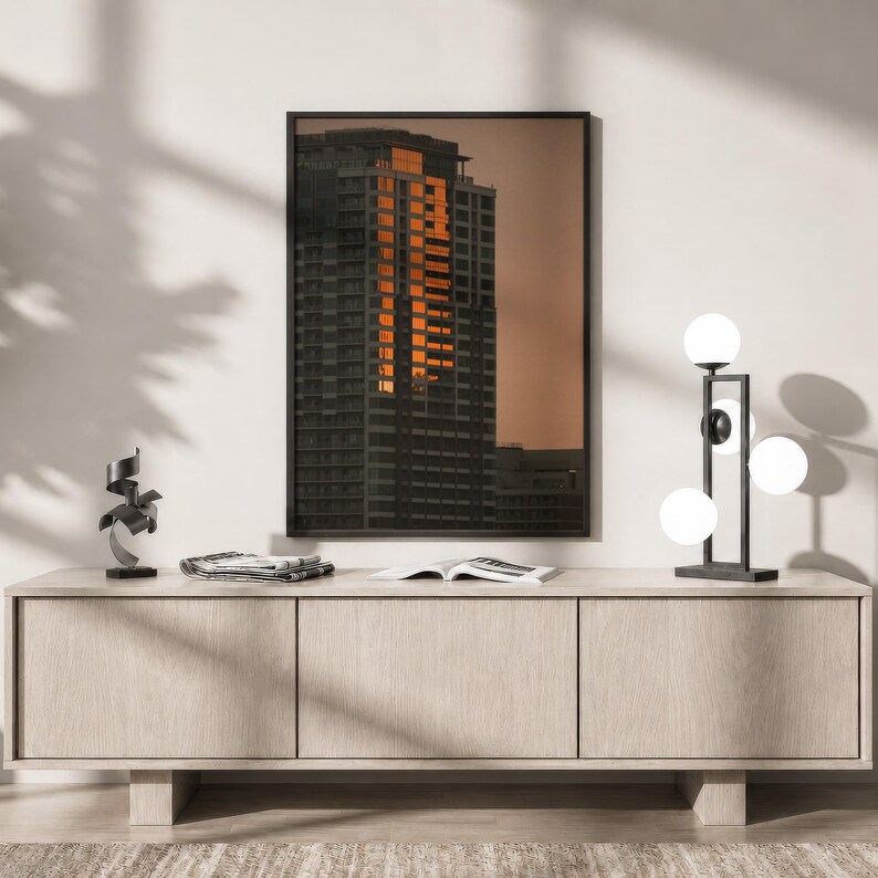 May include: A framed wall art print depicting a dark skyscraper with glowing orange windows at sunset, positioned above a light wood sideboard featuring a black metal abstract sculpture and a modern lamp with four white spherical glass globes.