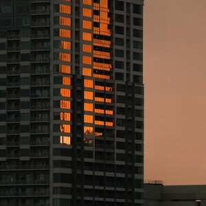 May include: A tall skyscraper with a grid of windows reflecting the bright orange light of a sunset. The building features numerous balconies and a dark gray facade, standing against a muted, dusky sky during the late evening hours.