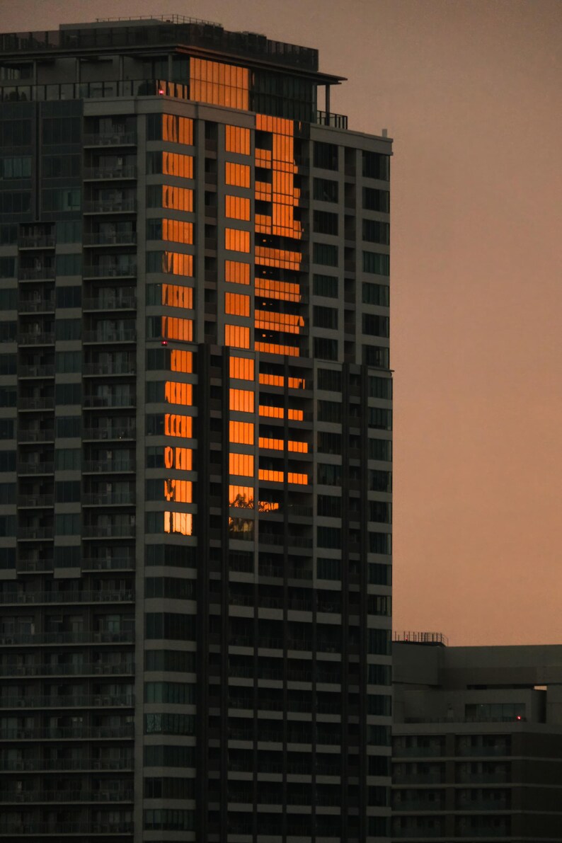 May include: A tall skyscraper with a grid of windows reflecting the bright orange light of a sunset. The building features numerous balconies and a dark gray facade, standing against a muted, dusky sky during the late evening hours.