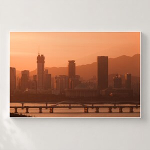 May include: A panoramic view of a city skyline at sunset, featuring multiple skyscrapers and a prominent domed building. A long bridge with a central arch spans across a river in the foreground, all silhouetted against a warm orange sky.