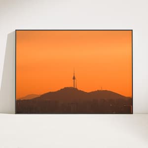 Puede incluir: Una impresi&oacute;n de arte mural enmarcada en formato horizontal que muestra la silueta de la torre N Seoul sobre una cresta monta&ntilde;osa contra un cielo naranja vibrante al atardecer. El primer plano muestra los contornos oscuros de una ciudad.