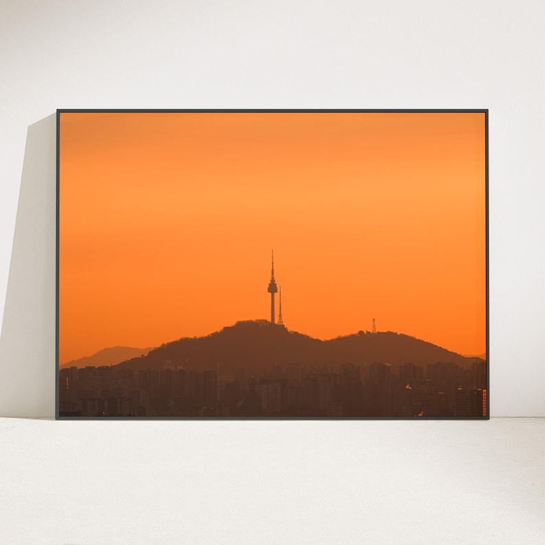Puede incluir: Una impresi&oacute;n de arte mural enmarcada en formato horizontal que muestra la silueta de la torre N Seoul sobre una cresta monta&ntilde;osa contra un cielo naranja vibrante al atardecer. El primer plano muestra los contornos oscuros de una ciudad.