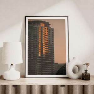 May include: A framed vertical wall art print featuring a high-rise building facade reflecting golden sunset light against a muted peach sky. The print is displayed on a wooden surface next to a white table lamp and a white ceramic donut vase.
