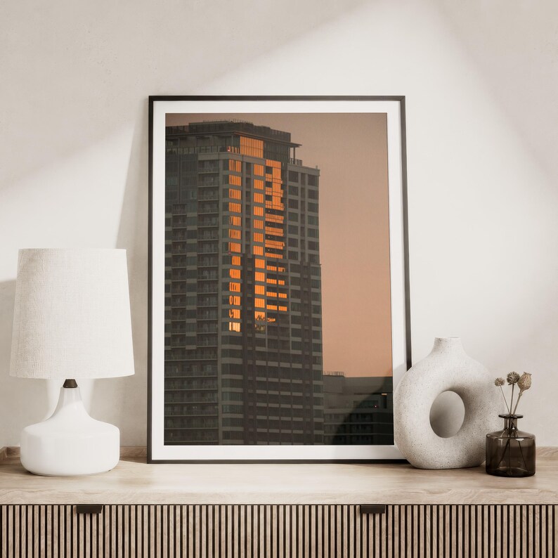 May include: A framed vertical wall art print featuring a high-rise building facade reflecting golden sunset light against a muted peach sky. The print is displayed on a wooden surface next to a white table lamp and a white ceramic donut vase.