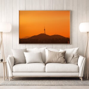 Puede incluir: Un cuadro rectangular que muestra la silueta de una alta torre de comunicaciones sobre una cadena monta&ntilde;osa contra un cielo naranja vibrante al atardecer, enmarcado en madera sobre un sof&aacute; blanco con varios cojines de tonos neutros.