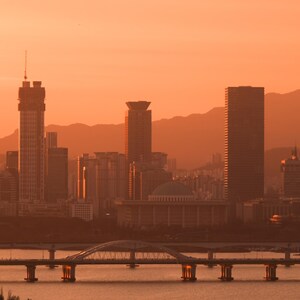May include: A cityscape featuring multiple skyscrapers and a domed building under a warm orange sunset sky. A long bridge with a central arch spans across the water in the foreground, with silhouettes of mountains visible in the distant background.