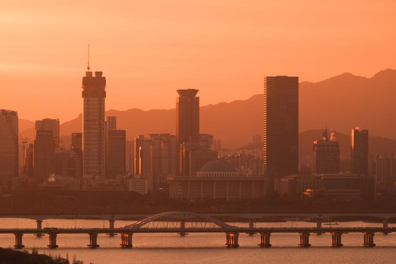 May include: A cityscape featuring multiple skyscrapers and a domed building under a warm orange sunset sky. A long bridge with a central arch spans across the water in the foreground, with silhouettes of mountains visible in the distant background.