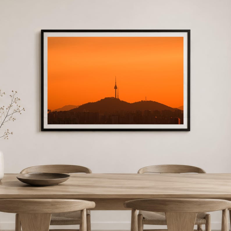 Puede incluir: Una impresi&oacute;n de arte mural de paisaje enmarcada que presenta la silueta de una torre de comunicaci&oacute;n alta sobre una cadena monta&ntilde;osa contra un cielo de atardecer naranja vibrante. La obra se exhibe en un marco negro delgado.