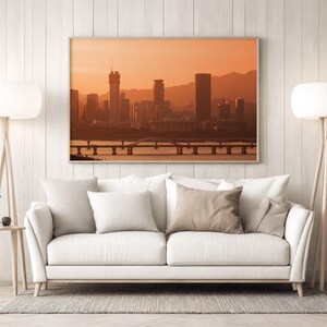 May include: A rectangular wall art print featuring a city skyline at sunset with orange hues, displayed above a white sofa with four throw pillows. The scene includes tall buildings, a bridge over water, and distant mountains in a warm colour palette.