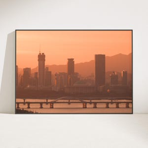 May include: A framed wall art print featuring a cityscape at sunset with a warm orange sky. The scene displays a river with a long bridge in the foreground, leading to a dense cluster of skyscrapers and a domed building against distant mountains.