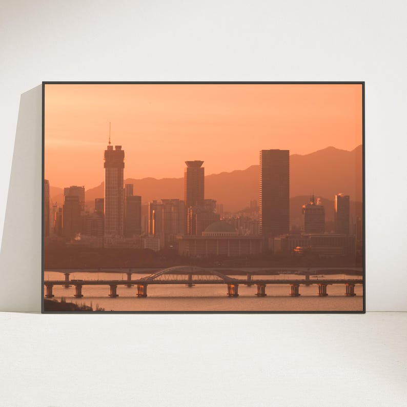 May include: A framed wall art print featuring a cityscape at sunset with a warm orange sky. The scene displays a river with a long bridge in the foreground, leading to a dense cluster of skyscrapers and a domed building against distant mountains.