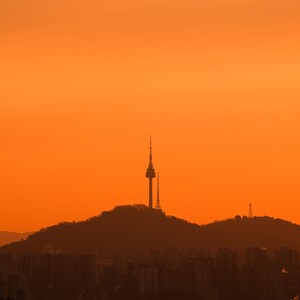 Puede incluir: Una silueta de la torre N Seoul sobre la cima de una monta&ntilde;a contra un cielo de atardecer naranja vibrante y s&oacute;lido. Los contornos oscuros de la torre y las colinas son visibles sobre la tenue silueta de una ciudad en el primer plano.